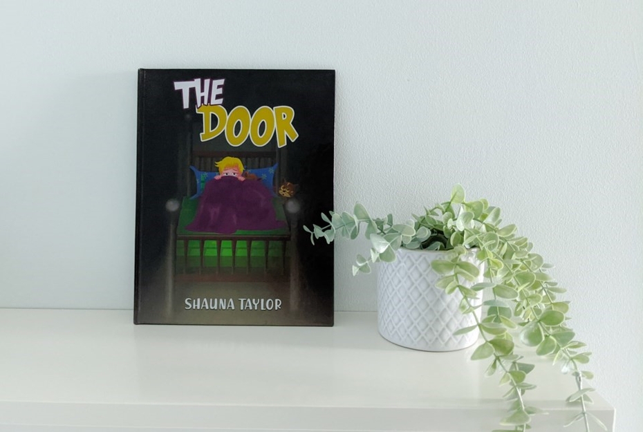 Children's book The Door by Shauna Taylor is displayed, leaning against a wall in a white, serene setting with a plant nearby.